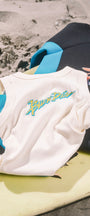 A white sleeveless shirt with a blue and green Buen Dia graphic, styled on a yellow surfboard with a black and blue wetsuit on a sandy beach background.