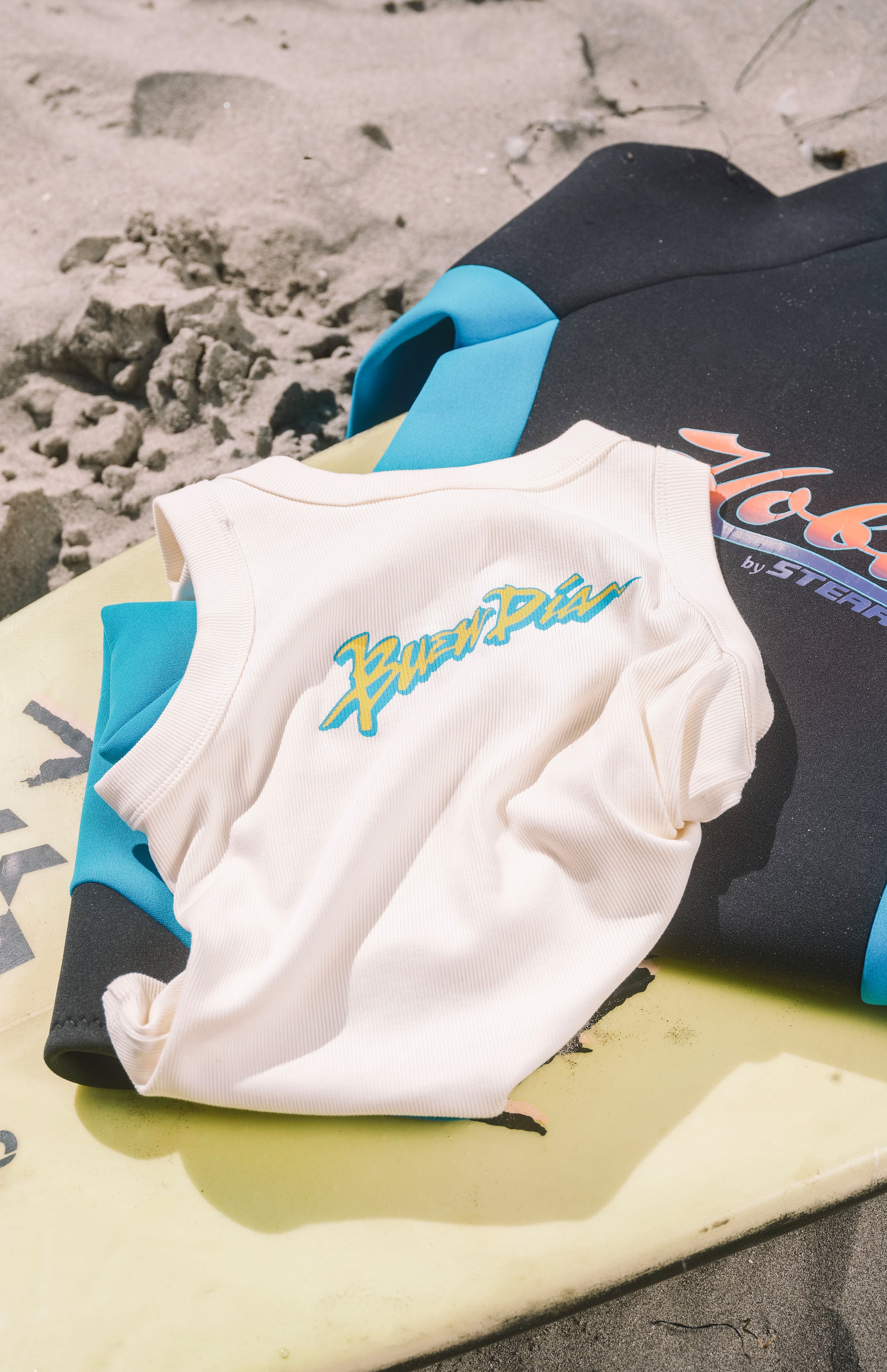 A white sleeveless shirt with a blue and green Buen Dia graphic, styled on a yellow surfboard with a black and blue wetsuit on a sandy beach background.