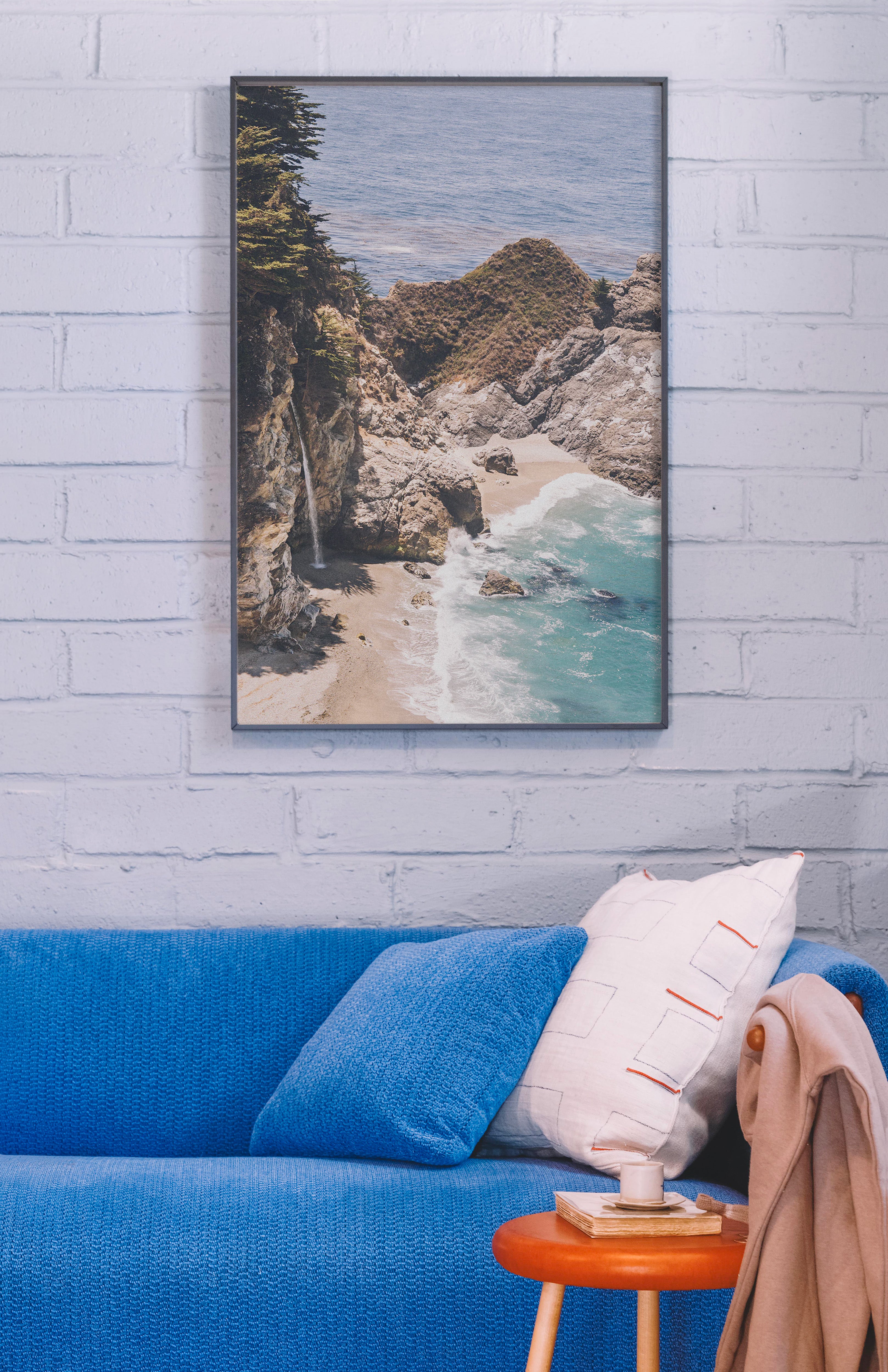 Framed beachscape print on a wall above a blue sofa with a white pillow. Buen Dia art prints San Luis Obispo