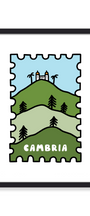 A framed art print featuring a stylized postage stamp design with rolling green hills, palm trees, a small white building on top, and the word 'CAMBRIA' in white. The print is mounted with a wide white mat and enclosed in a slim black frame against a light gray background.