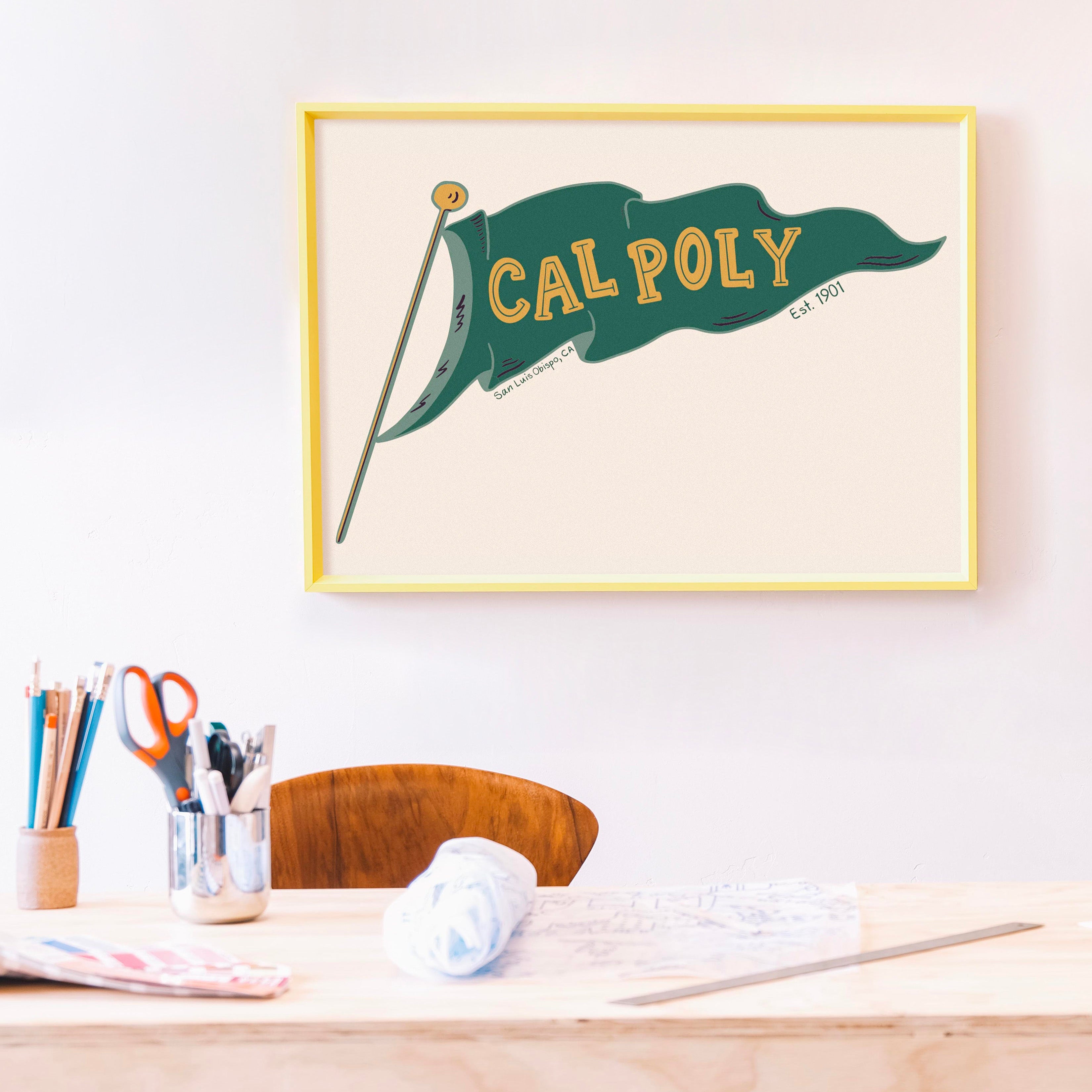 Framed artwork with CAL POLY flag on a wall above a desk.