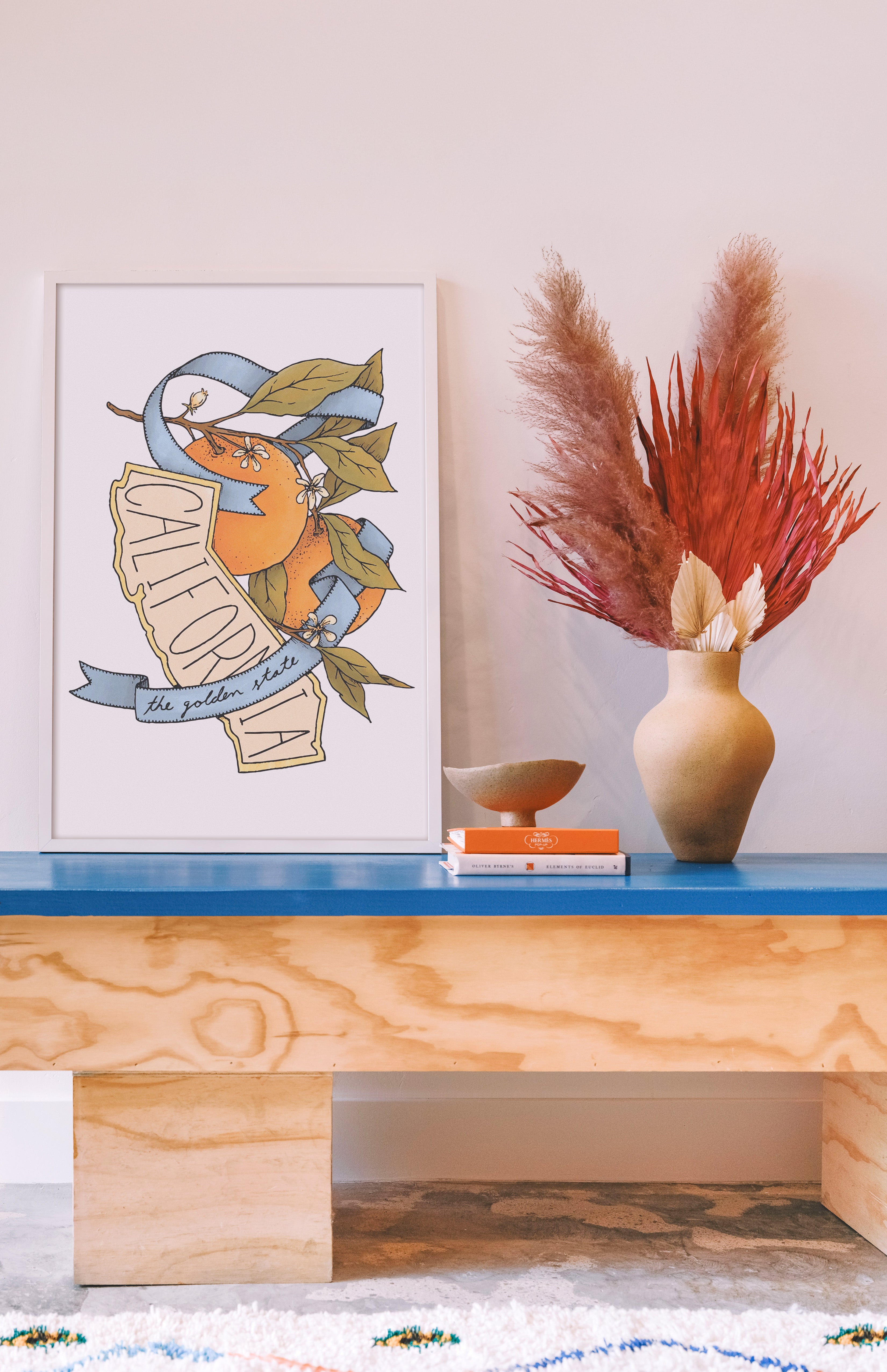 Wooden console table with decorative items including a vase, books, and a framed print on a light-colored wall. Buen Dia art prints San Luis Obispo