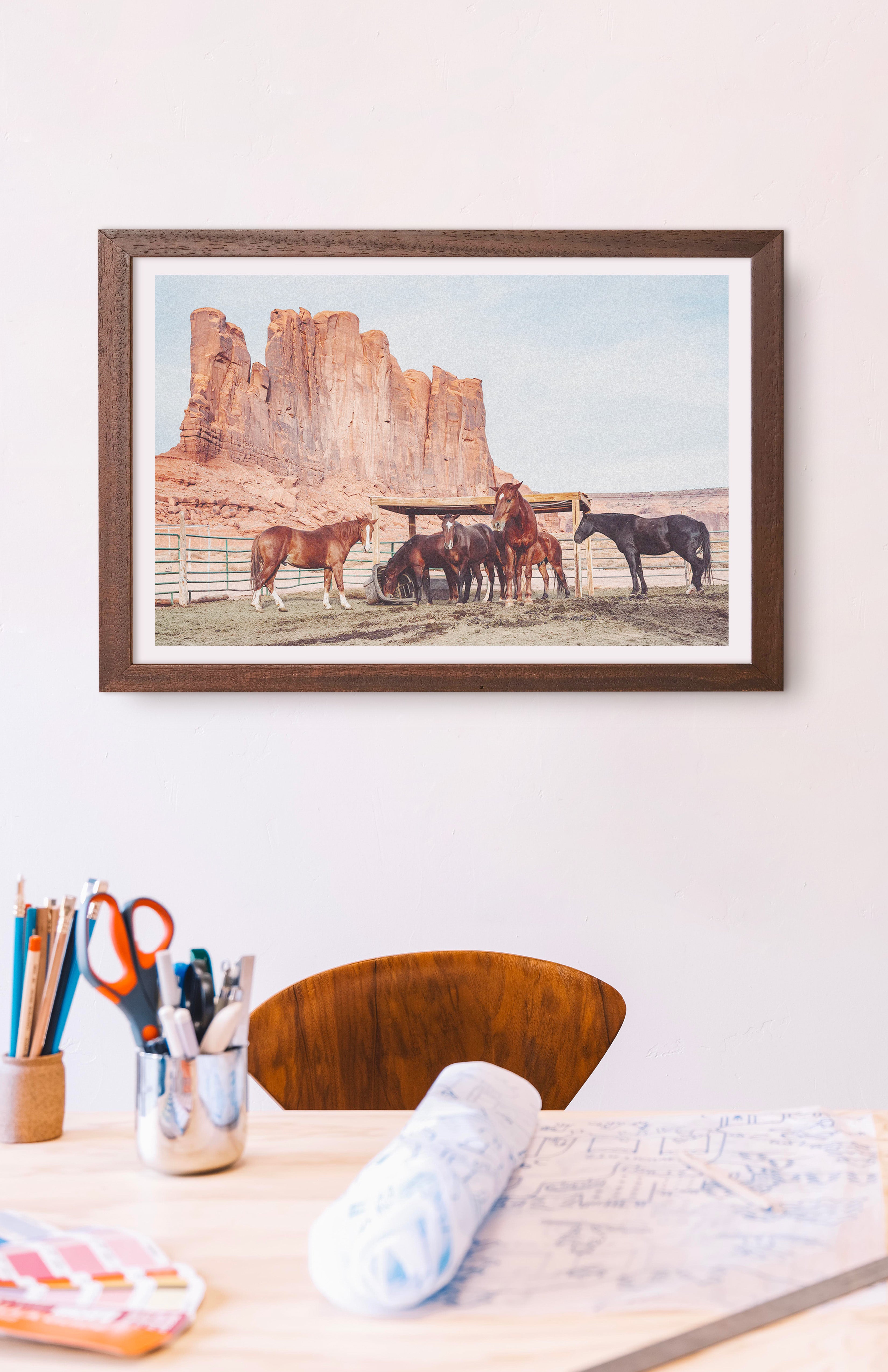 Framed artwork of horses in a desert landscape above a desk with office supplies. Buen Dia art prints San Luis Obispo