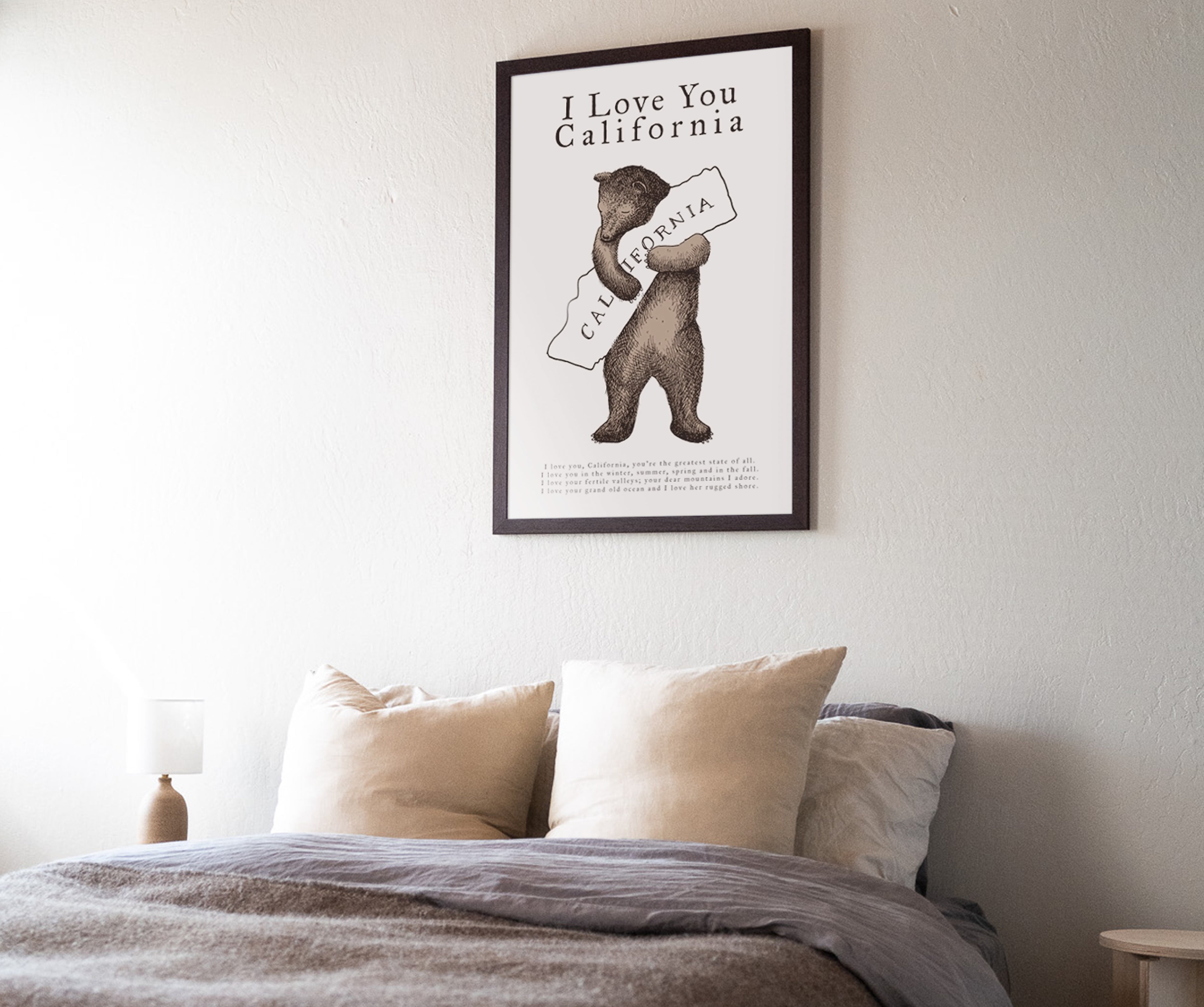Framed poster of a bear holding 'California' above a bed with pillows and bedding. Buen Dia art prints San Luis Obispo.