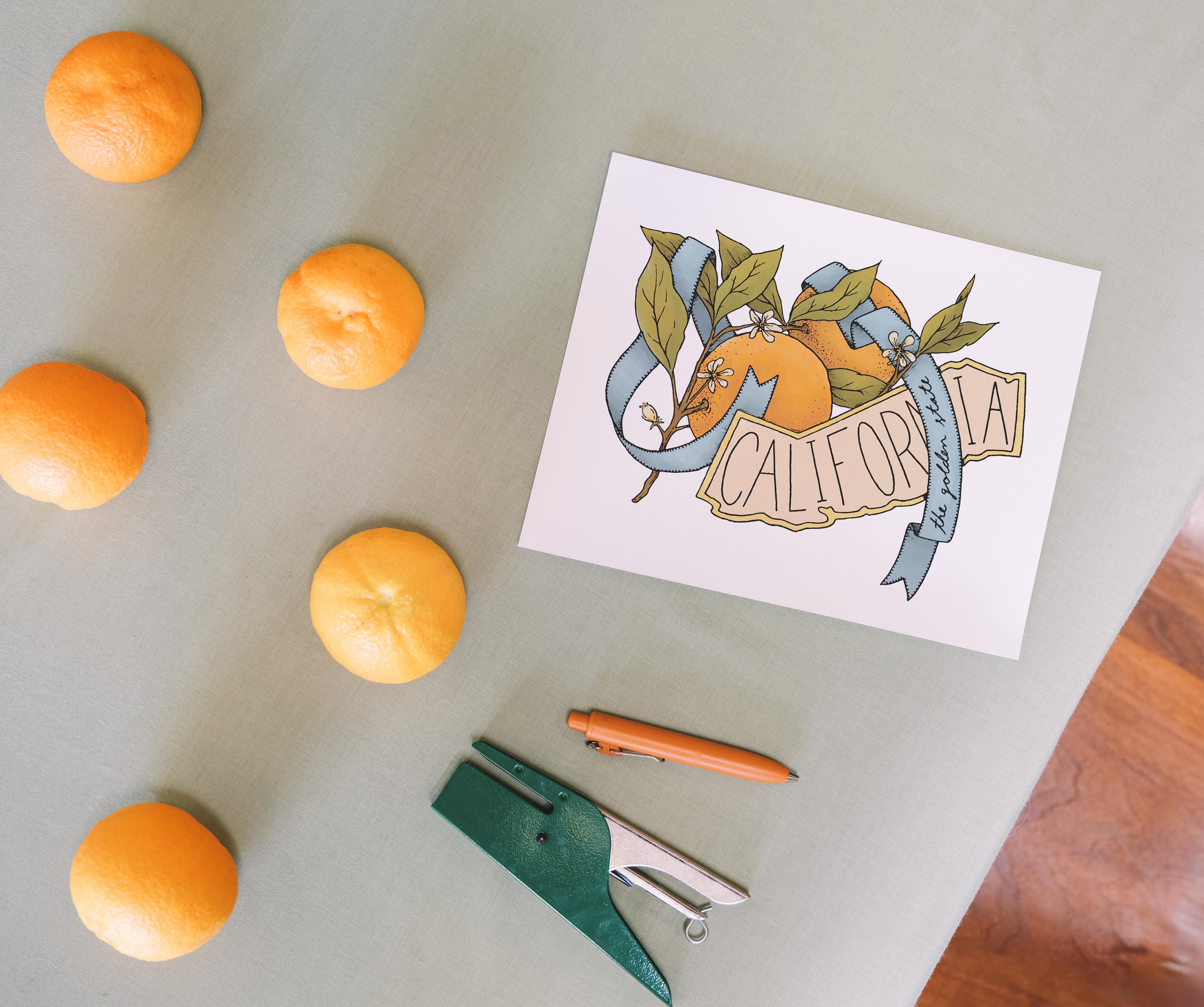 Art Print of California state, surrounded by oranges and a stapler on a light surface. Buen Dia art prints San Luis Obispo.