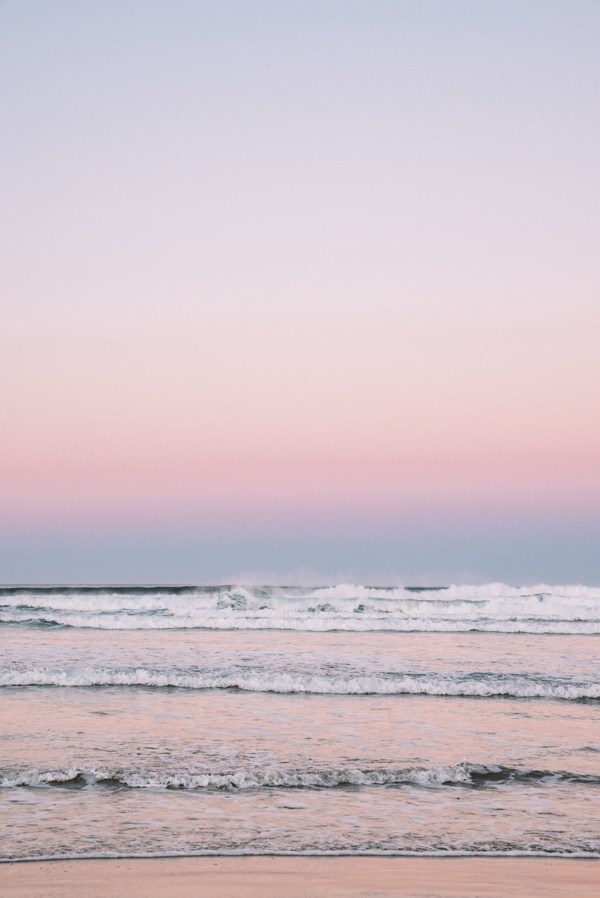 Pastel pink and blue gradient sky over a calm ocean with a sandy beach.